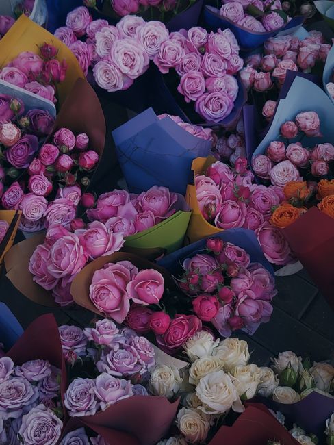 A variety of freshly cut roses and peonies in shades of pink, purple, white, and orange arranged in colorful wrapping paper and paper cones. The flowers have delicate, layered petals with visible text