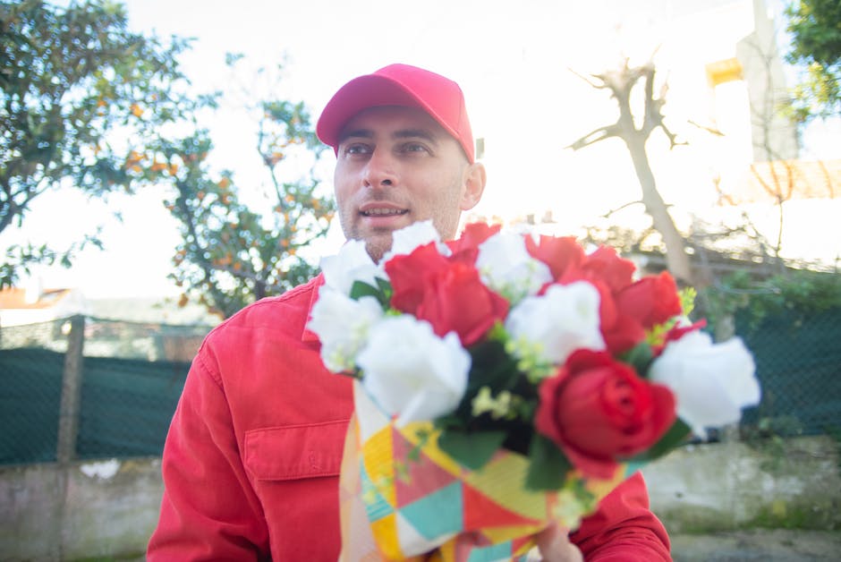 A man in a red cap and matching red jacket holds out a vibrant floral bouquet towards the camera. The bouquet features a mix of red and white blooms, including roses and other fresh flowers, arranged