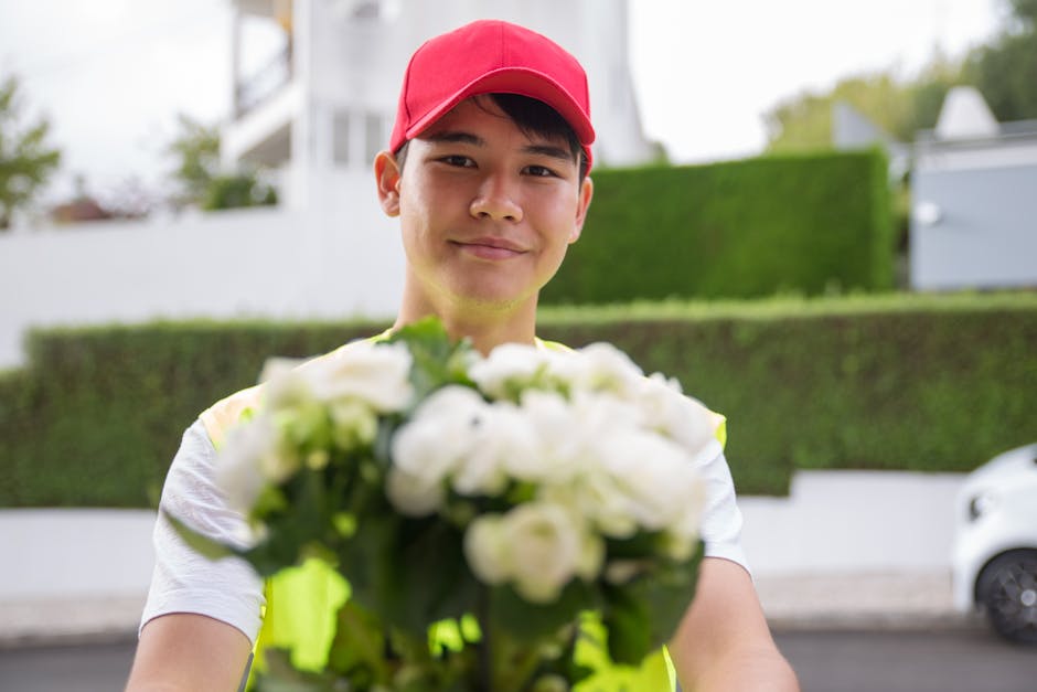A young male flower delivery courier wearing a red cap and a high-visibility vest, smiling and holding a bouquet of white roses and greenery towards the camera. The bouquet is wrapped in clear plastic
