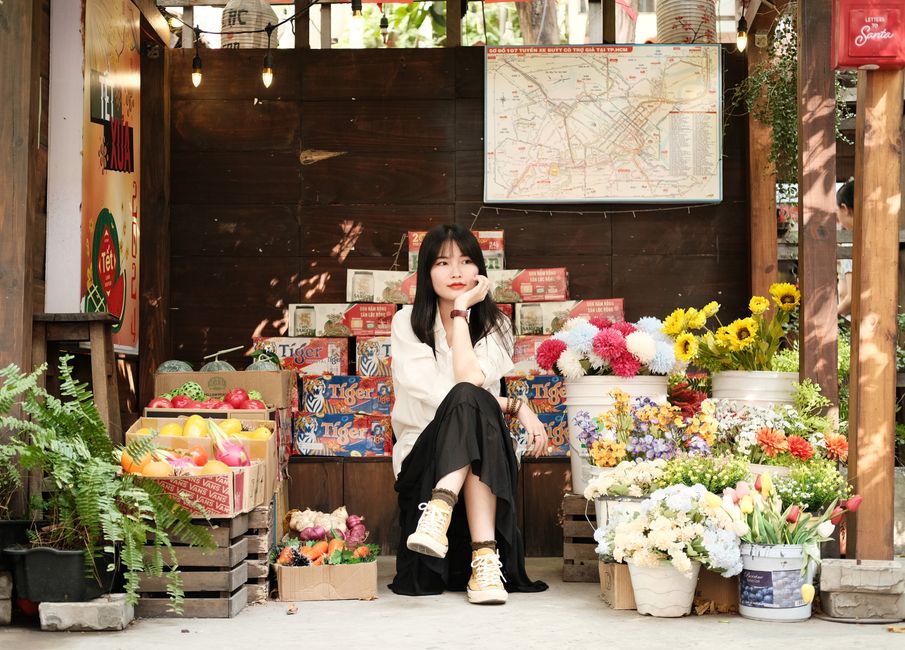 A young woman with long dark hair, dressed in a white top and black skirt, sits on a low wooden platform amid a vibrant flower market display. She rests her chin on her hand, gazing thoughtfully. Surr