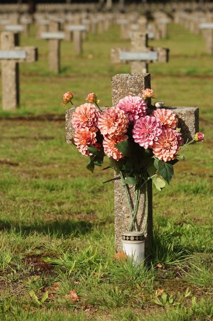 A close-up of a floral arrangement attached to a weathered, grey stone cross in a churchyard or memorial garden. The bouquet features a cluster of large, soft pink and peach dahlias with ruffled petal